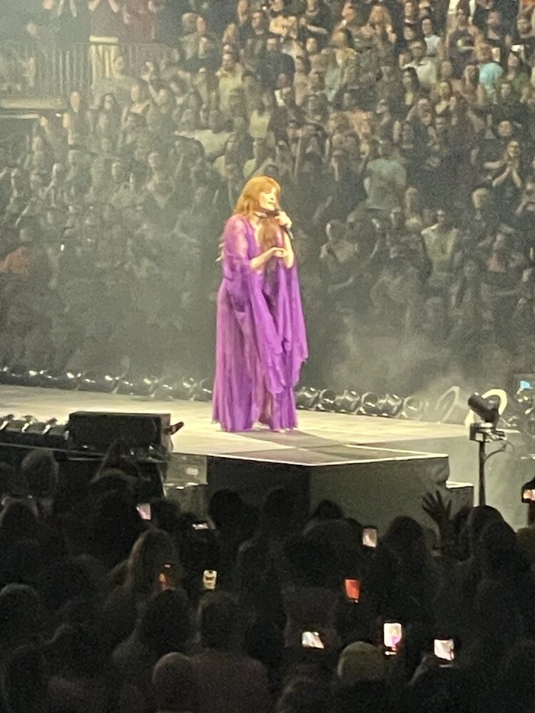 A stadium audience jumping with hands raised, moving together at a Florence + the Machine show